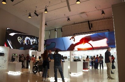 Trade show floor with illuminated product kiosks, attendees chatting in small groups, and a large digital display showing EV charging equipment overhead.
